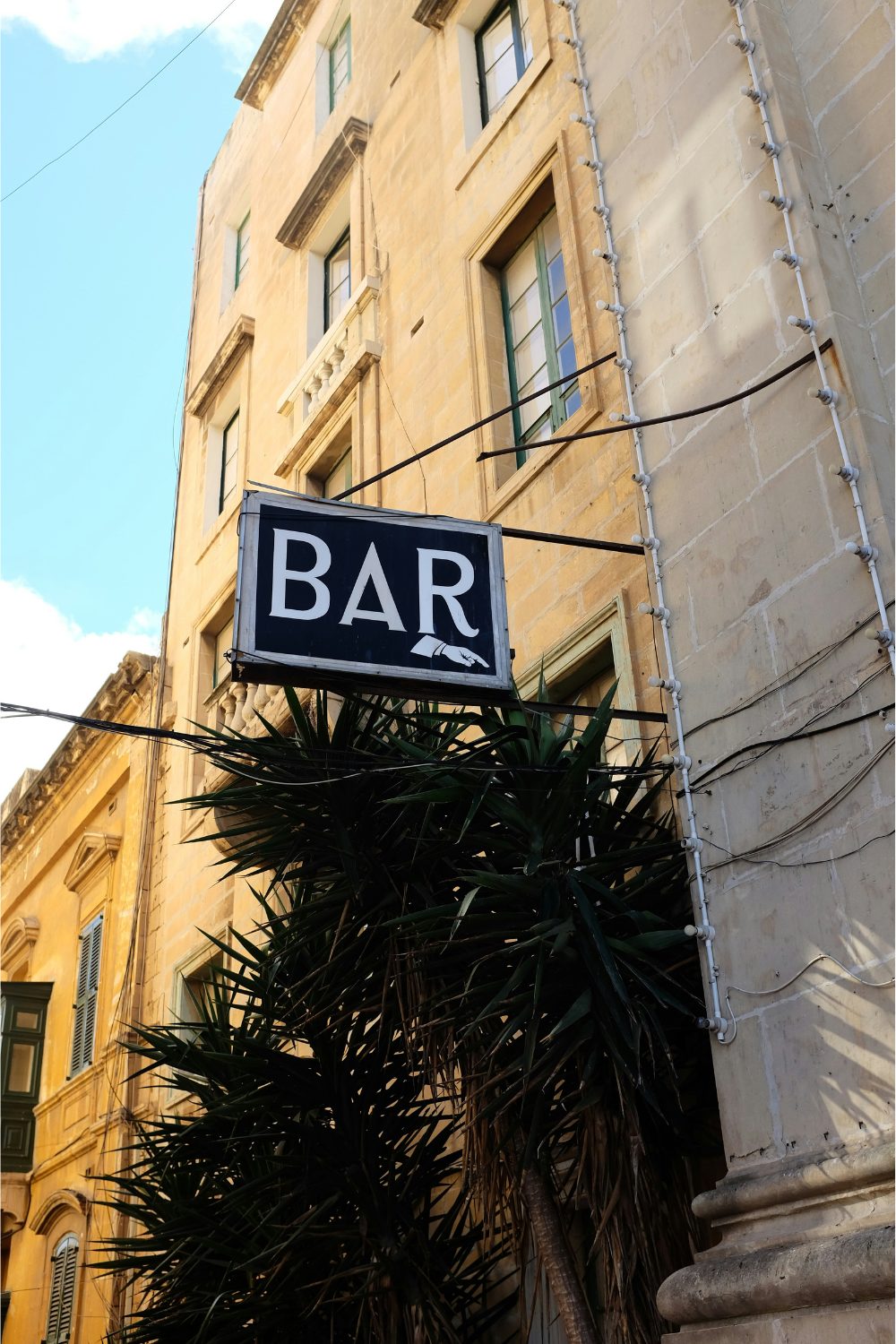 Exterior of yellow stone building with black and white Bar sign above spiky plants
