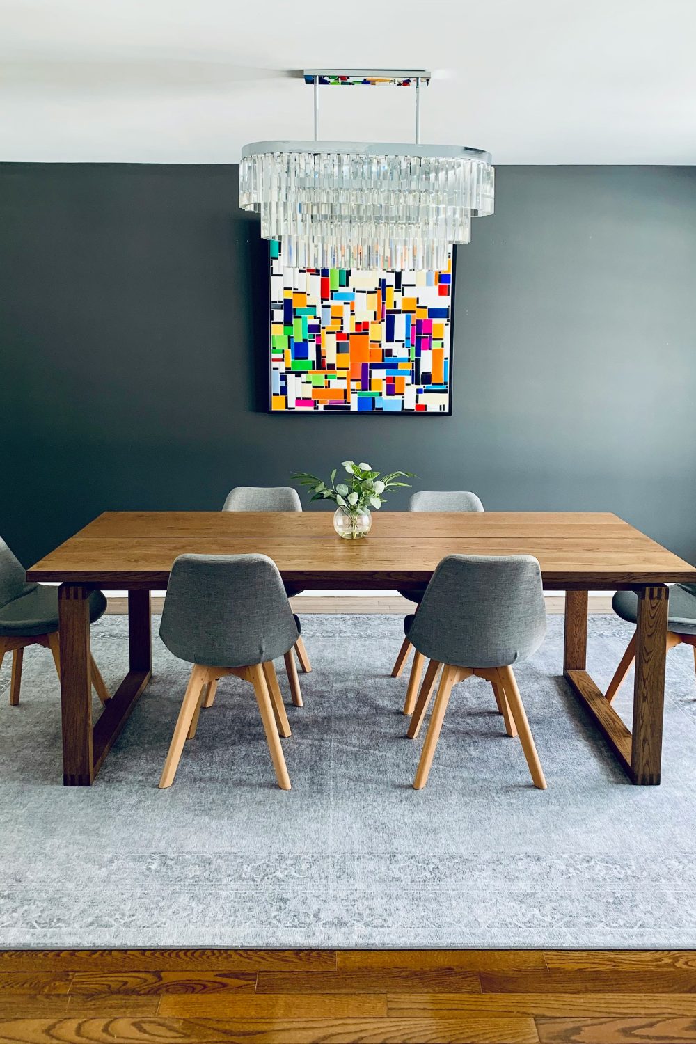 Wooden dining table with grey chairs under glass oval chandelier and colourful abstract print on a grey wall