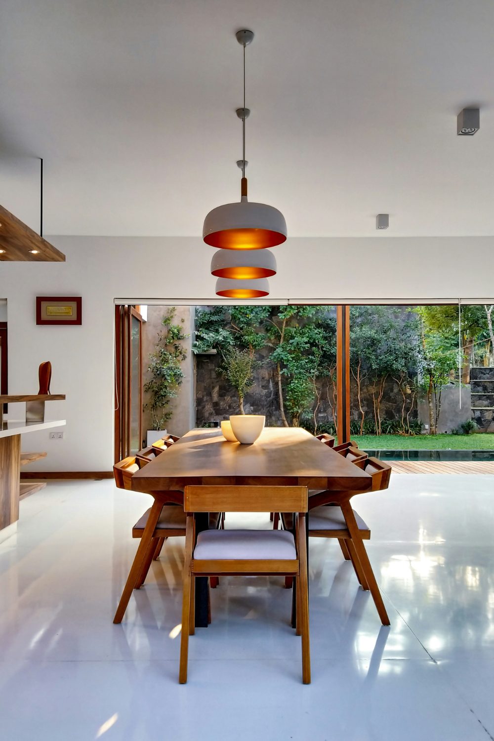 Wooden dining table and chairs under three metal lamps and a garden view through French windows