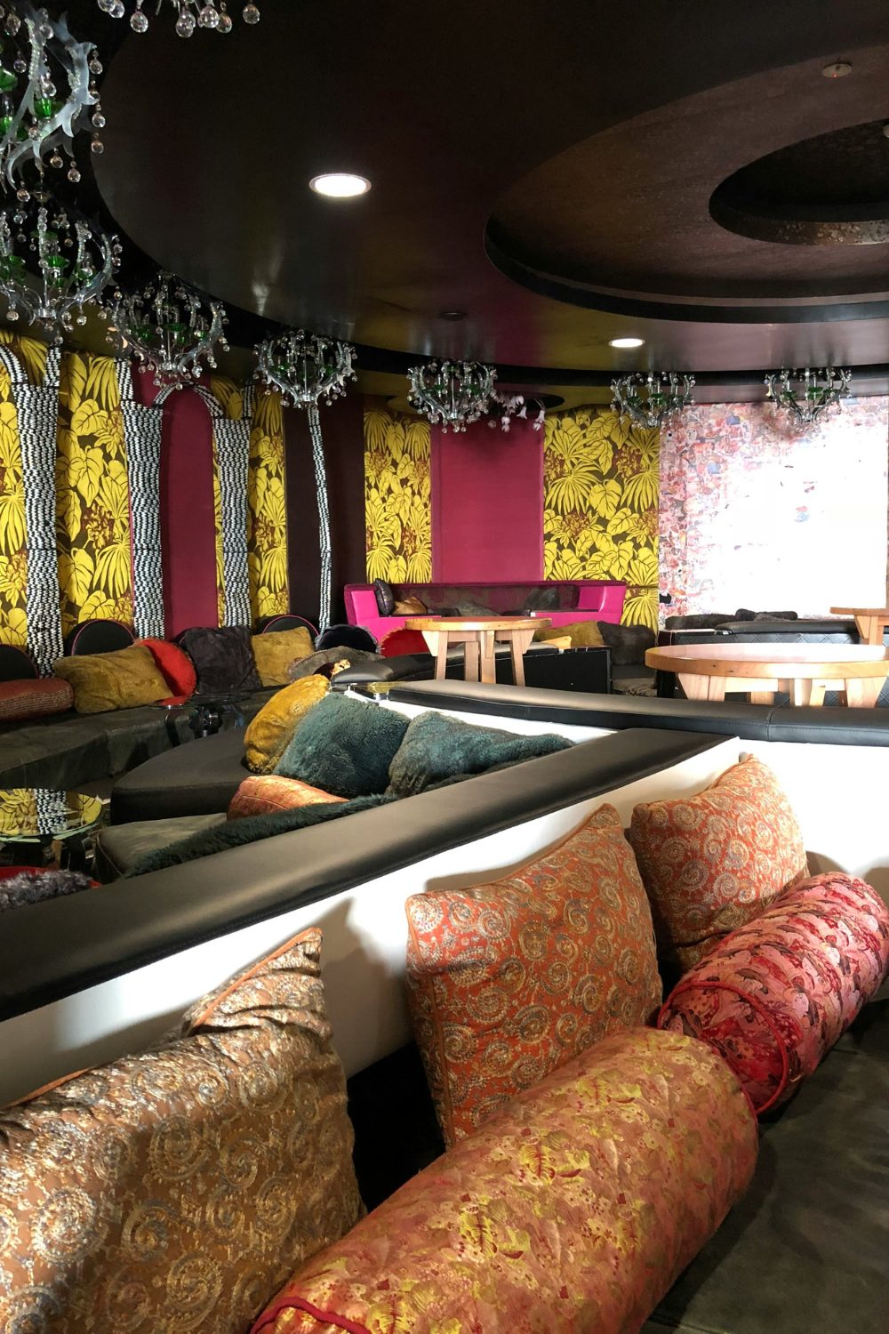 Bright cafe interior with chandeliers, sofas with luxurious colourful cushions, and yellow plant print wallpaper