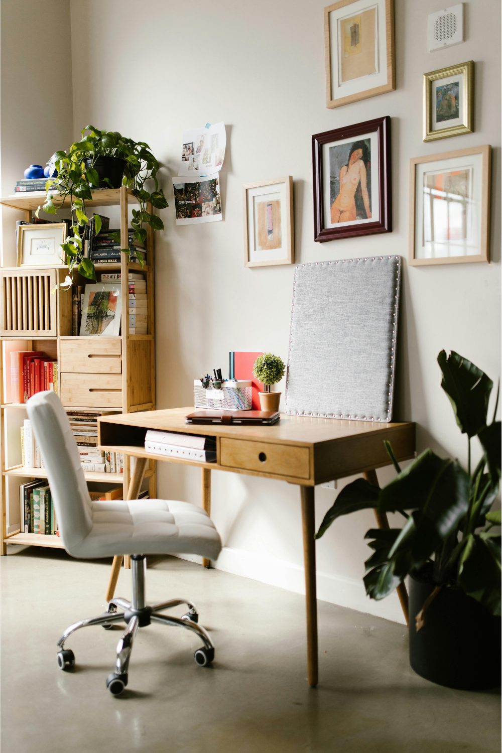 Wooden desk with grey office chair next to bookshelf and framed prints on white wall
