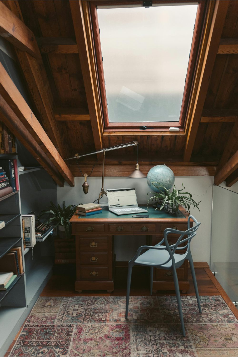 Home office in attic space with wooden desk, grey chair and faded rug below large window