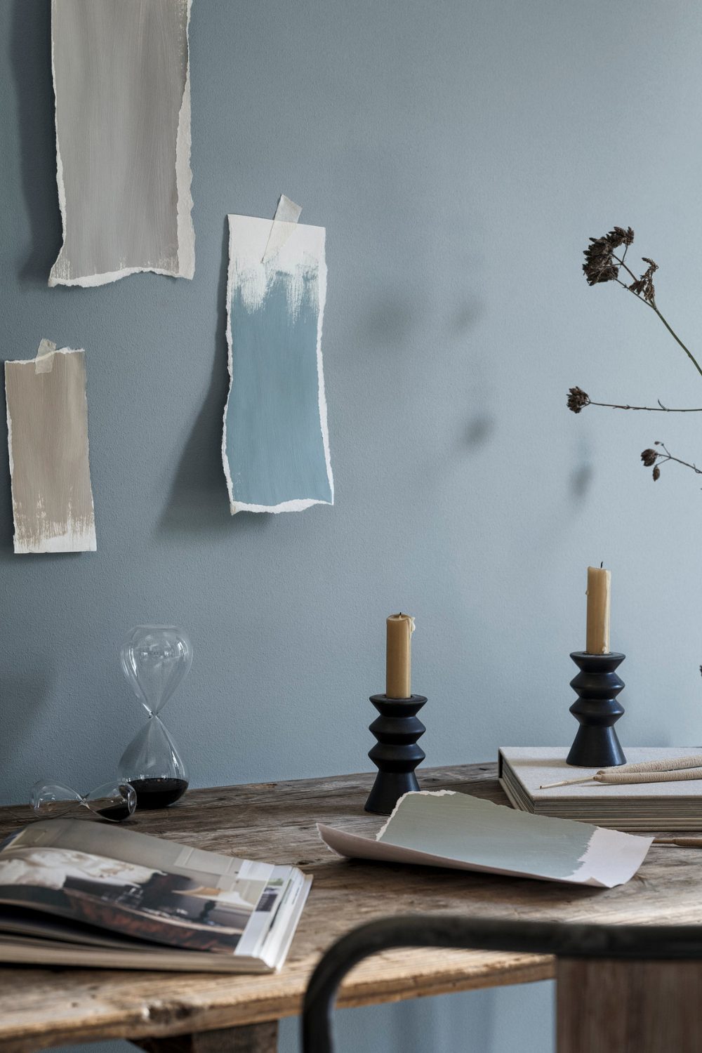 Paint swatches taped to a dusky blue wall above a wooden desk with candlesticks and hourglass