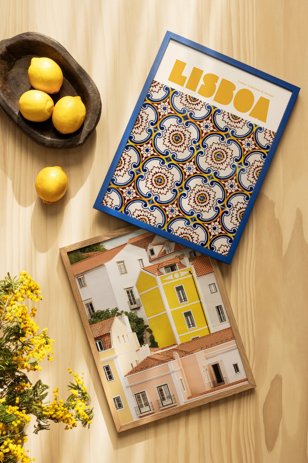 Two framed prints of Lisbon travel posters on wooden table next to black bowl of lemons and yellow flowers
