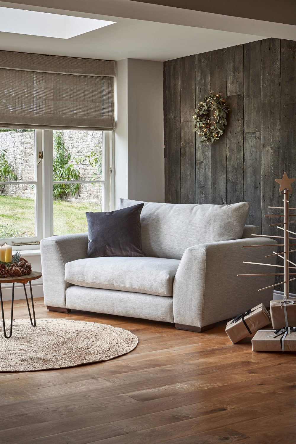 Grey loveseat with circular jute rug in front of wooden wall and next to window with garden view