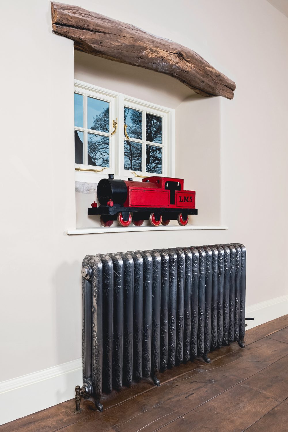 An ornate silver radiator below a window with a red model train and natural wooden lintel