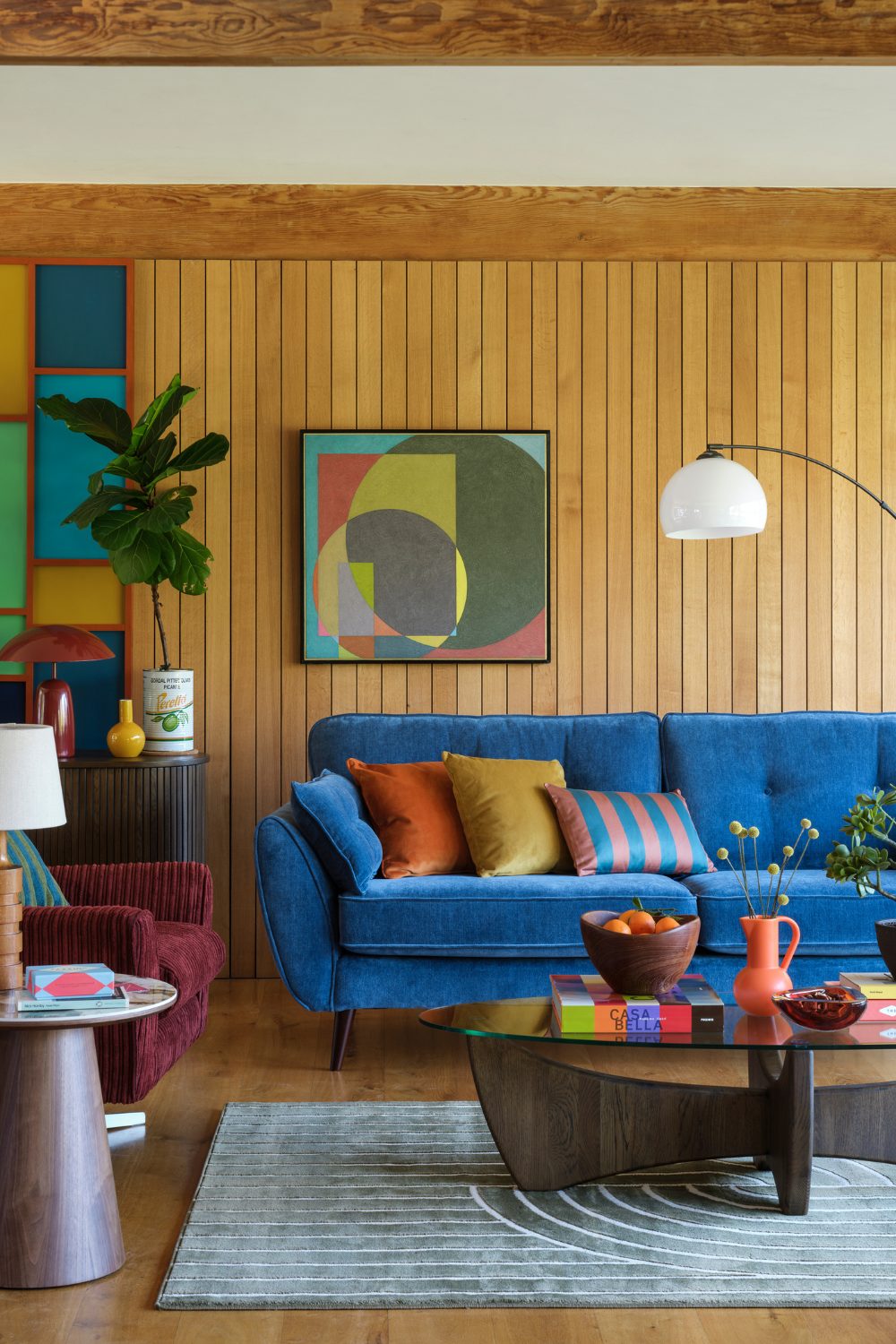Blue sofa in colourful living room with wood panelled wall, red chair and framed multicoloured print