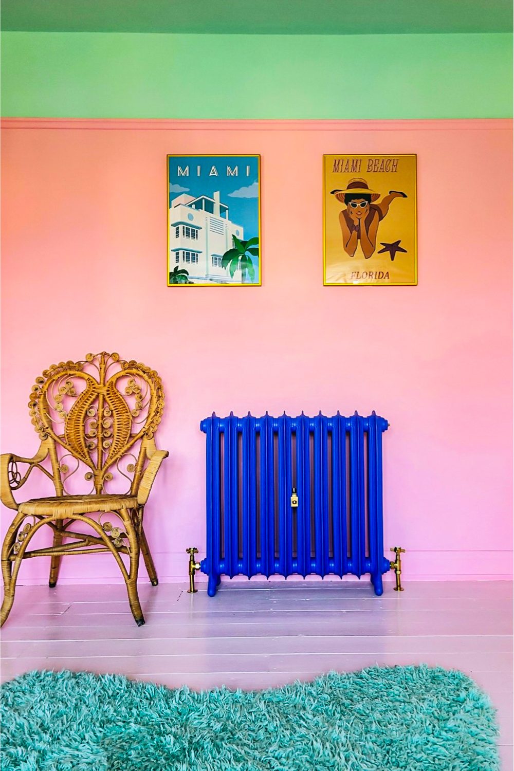 Royal blue radiator on an ombre pink wall next to an ornate gold wicker chair. Vintage travel posters are on the wall and a turquoise rug is in the foreground.