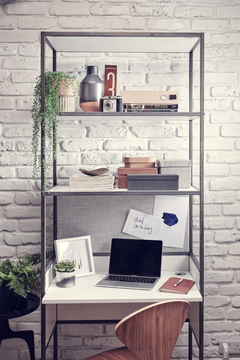 Black metal shelving unit holding boxes, notebooks and houseplants with built-in desk and wooden chair