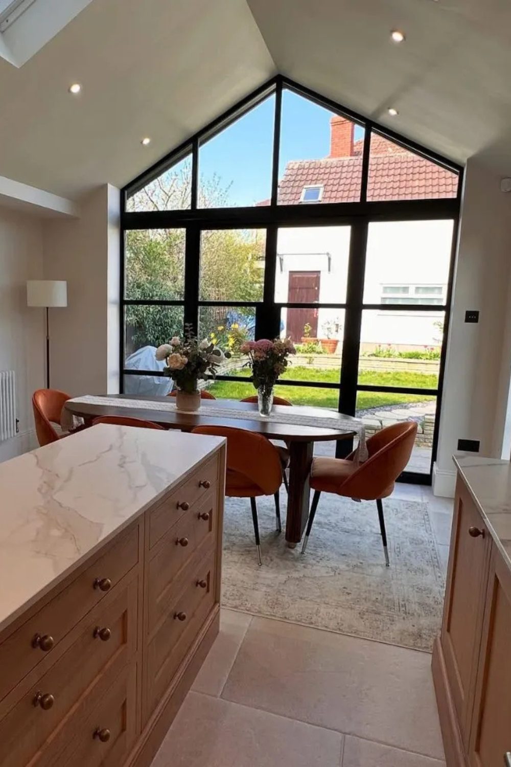 Kitchen with glass wall and garden view, wooden table with orange chairs, and marble and wood counters