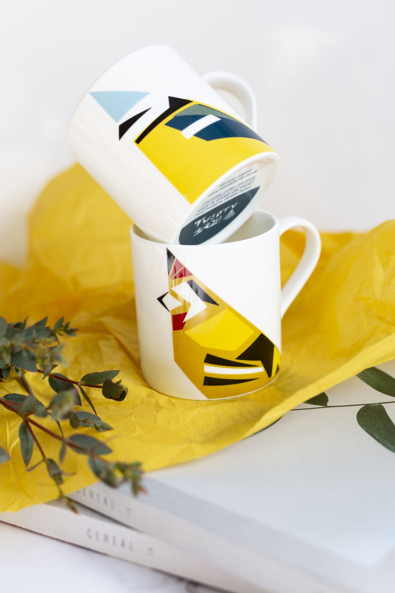 Two white mugs with stylised cubist birds designs on yellow tissue paper and stack of white books