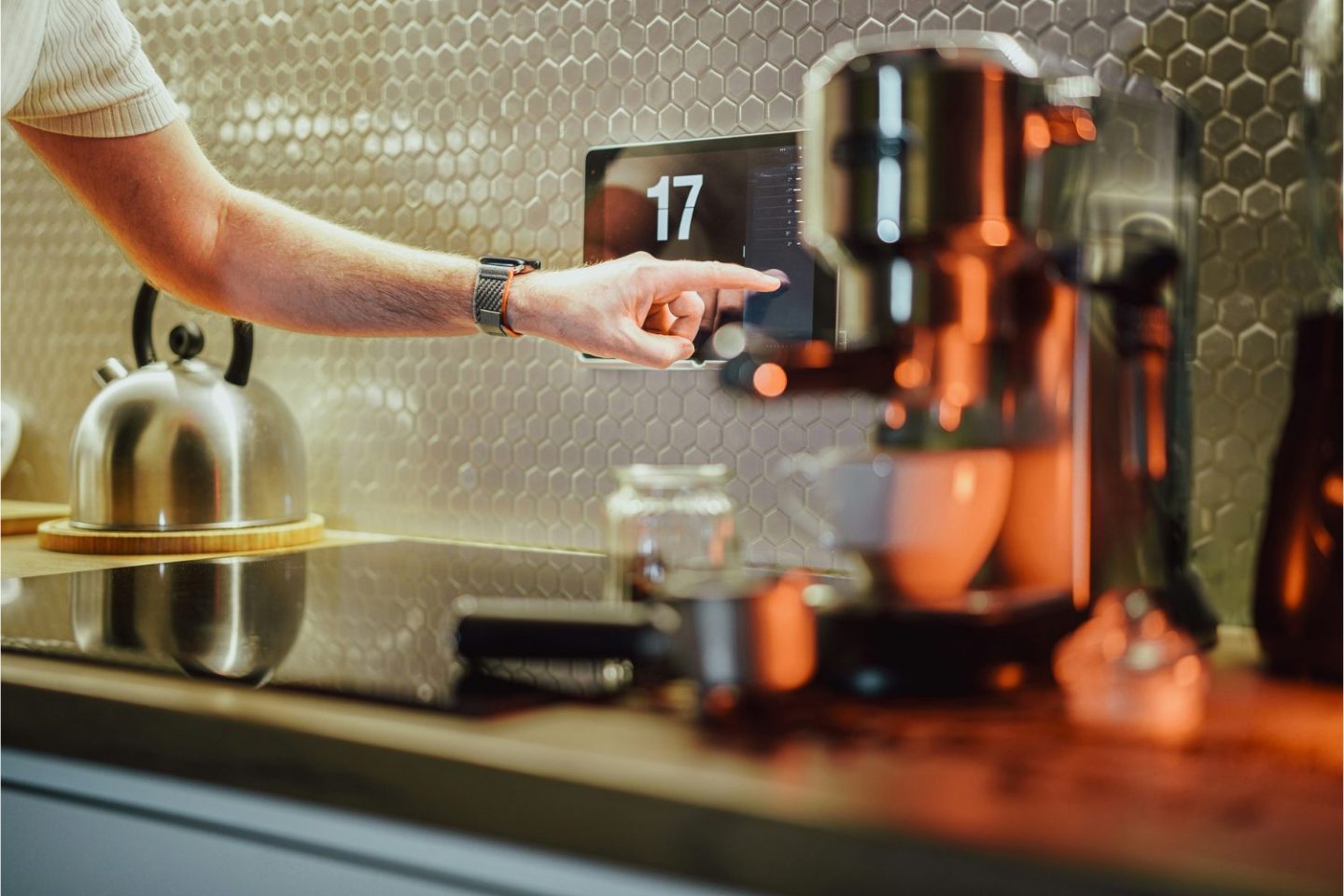 Someone pushes a button on a smart thermostat in a white tiled kitchen with kettle and coffee machine