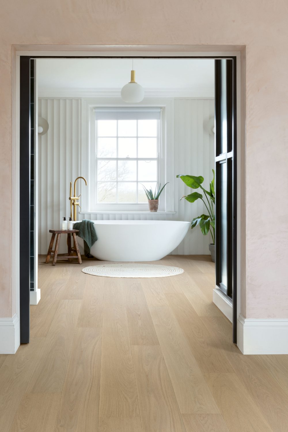 Freestanding white bath on wooden flooring in a bathroom with fluted white wall and pot plants