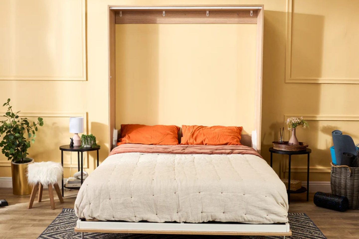 Murphy bed with orange pillows in yellow room next to bedside tables, gold plant pot and wicker basket.