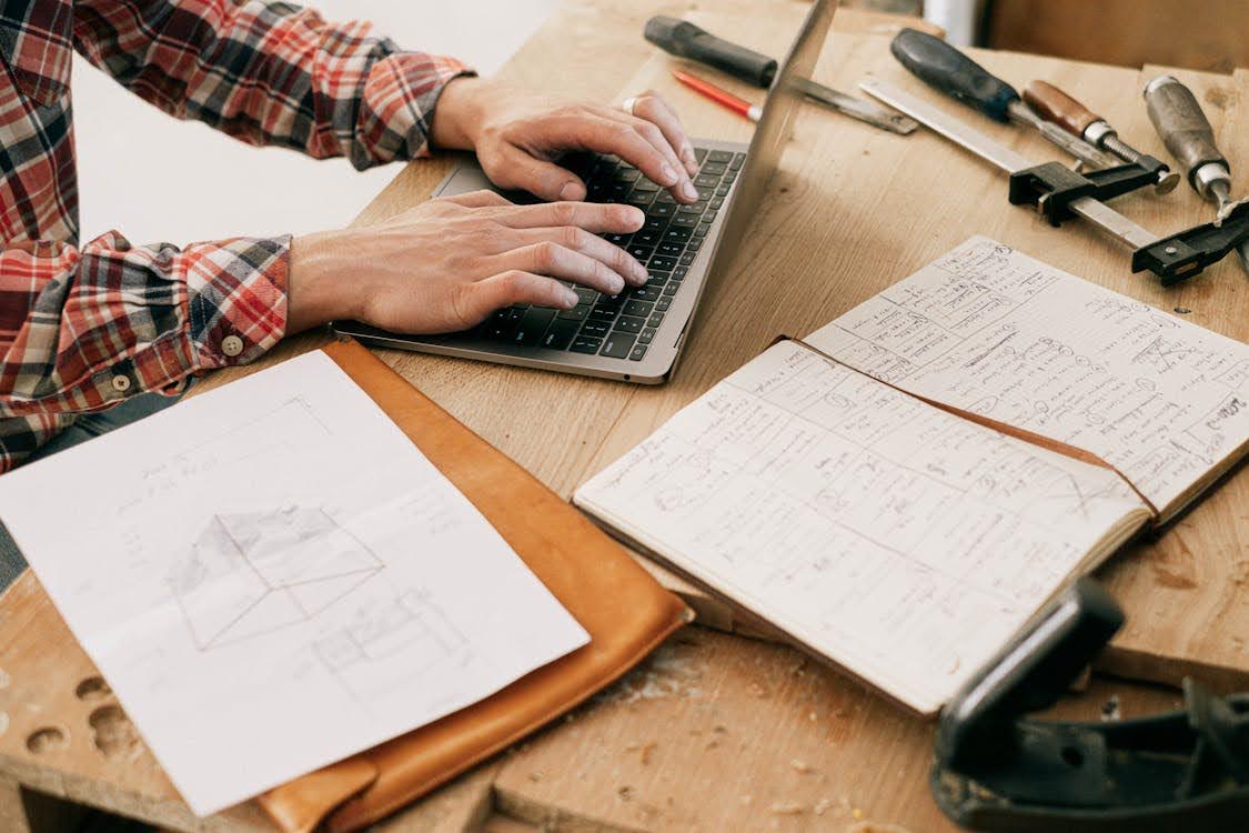 A man's hands working on a laptop on a wooden desk surrounded by tools and notebook