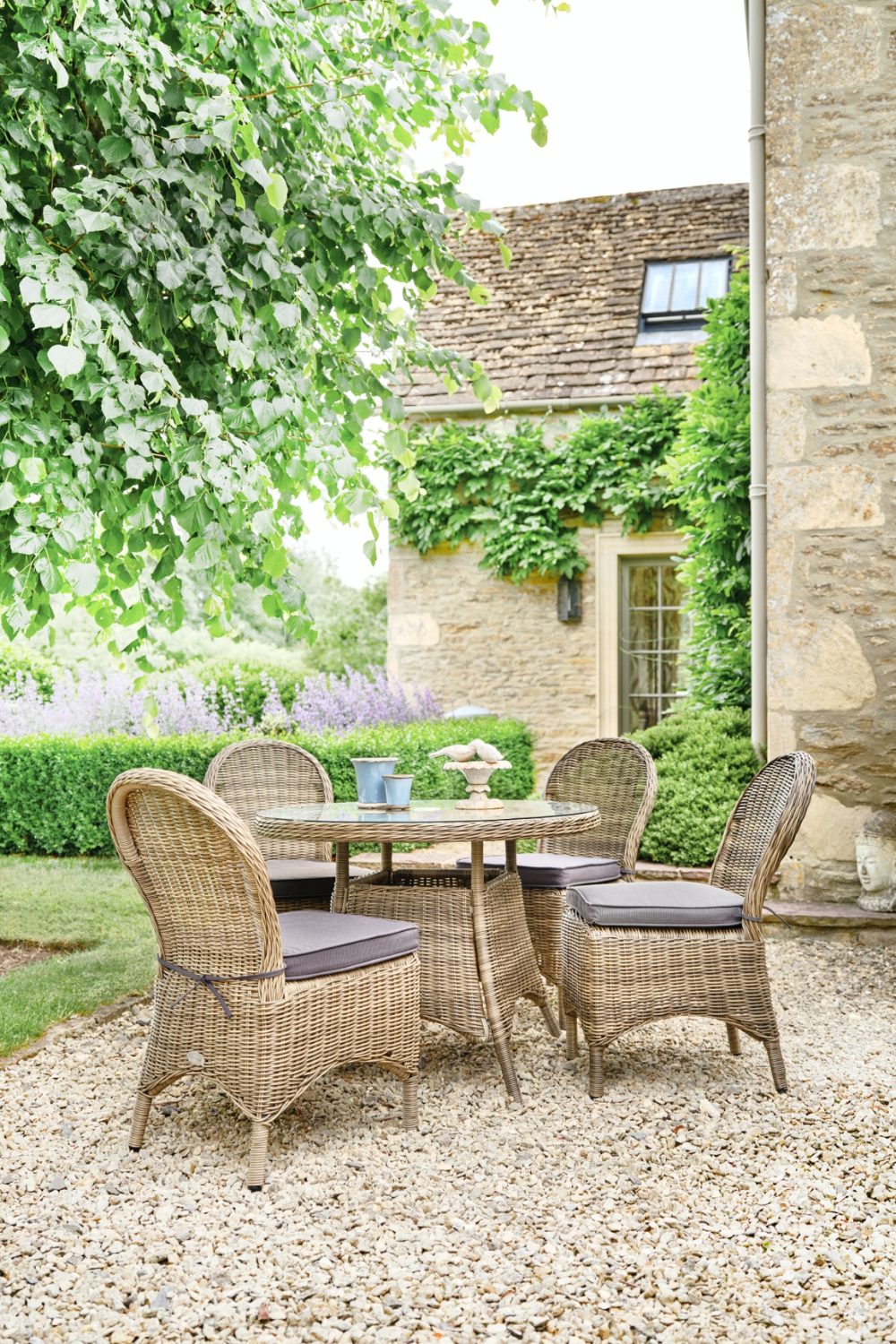 Wicker table and chair set on gravel in a garden with low hedge and purple flowers in front of stone house