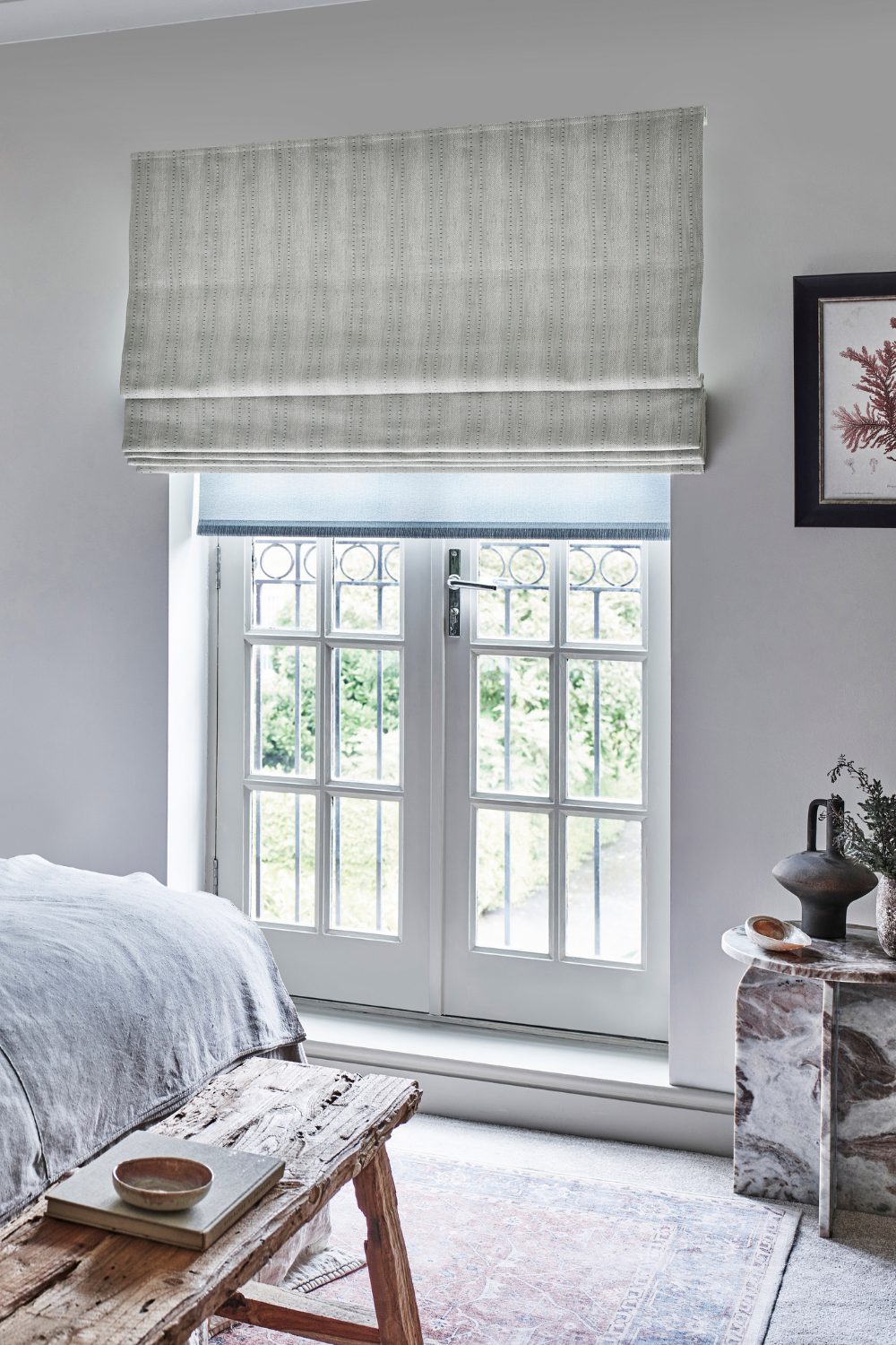 White bedroom with white French doors, distressed wooden bench, marble table, and white striped blind