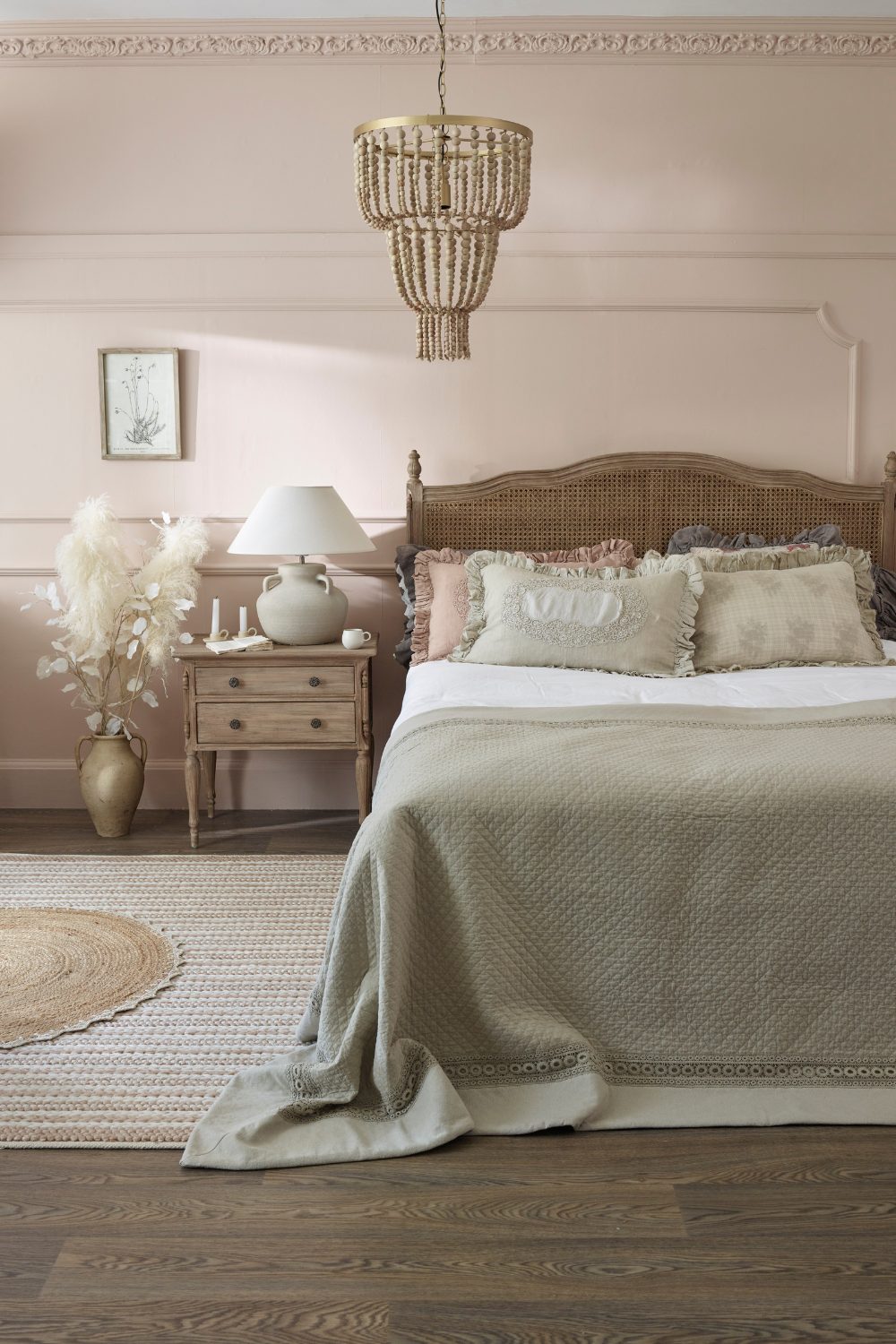 Light pink bedroom with wooden headboard, wooden bedside drawers, circular jute rug, wooden chandelier and frilled pillows