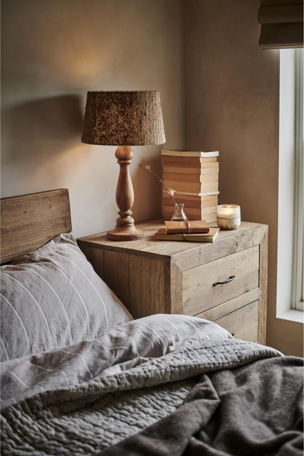 Wooden bedside table with candle, lamp and book stack, next to wooden bed with grey bedding