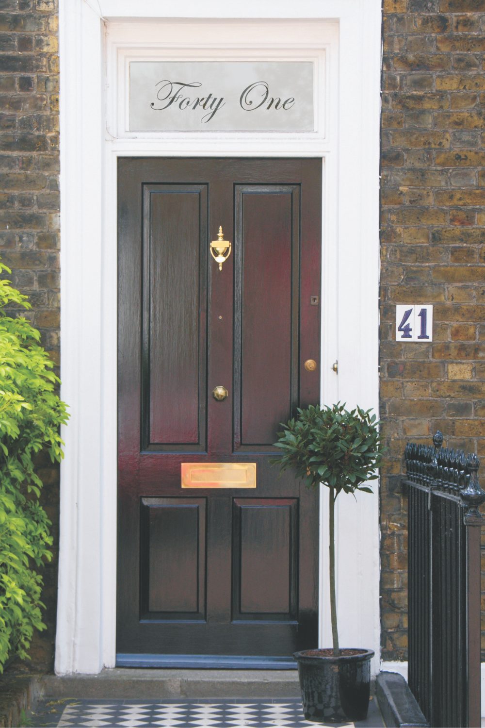 Black front door in brick house with patterned black and white path and potted tree