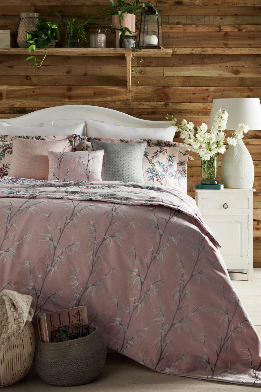 Floral bed next to white drawers holding white lamp and plant pot in front of wooden wall with high shelf
