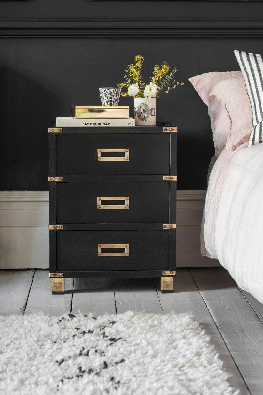 Black drawers with gold fittings holding books and plant pot next to bed with striped pillow