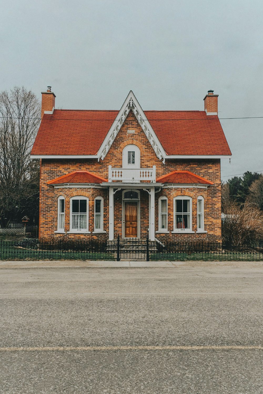 Red brick house with red roof, white balcony and peaked gable behind wide road against grey sky