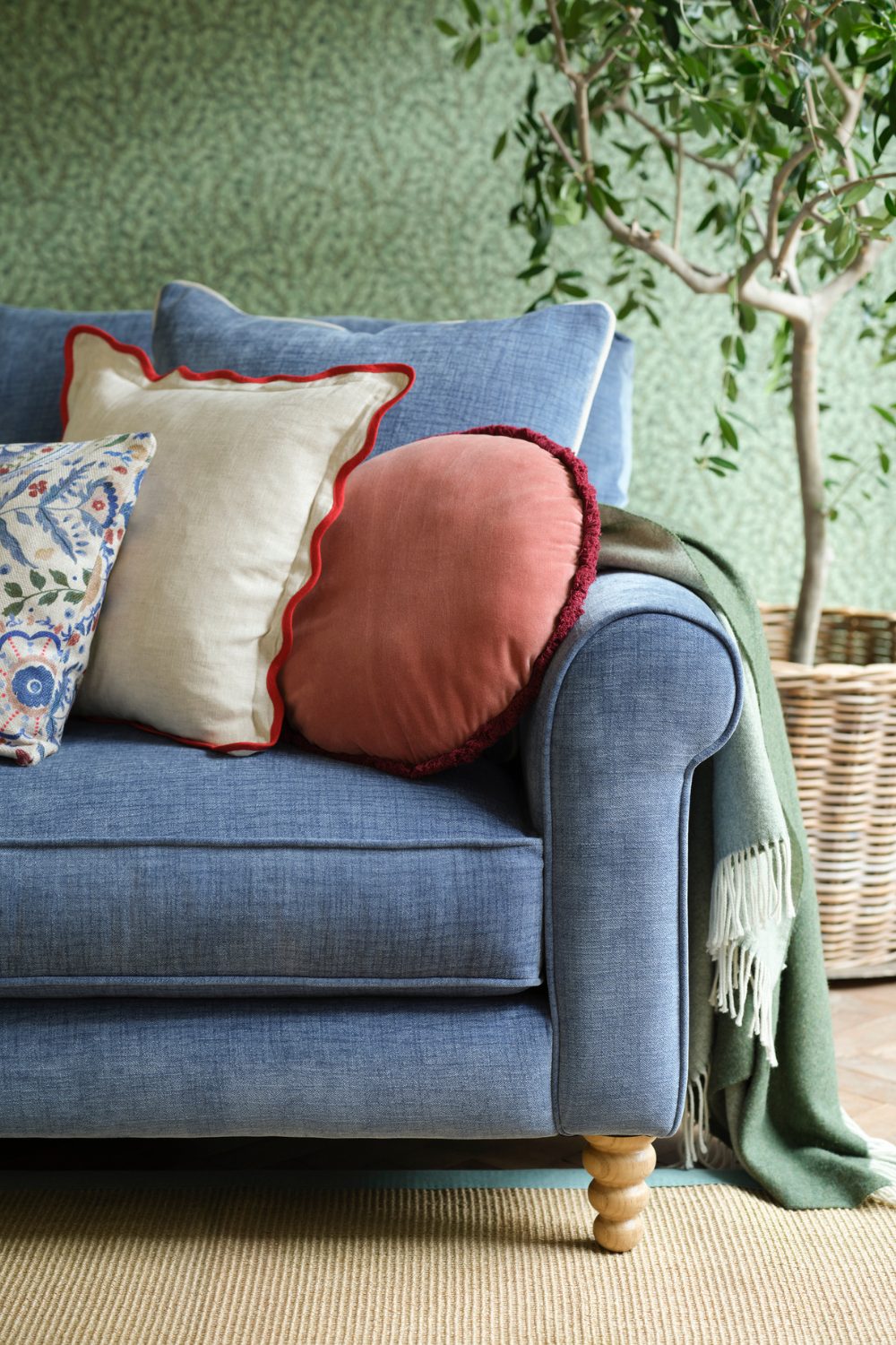 Blue sofa with three assorted throw pillows next to small potted tree against green patterned wallpaper