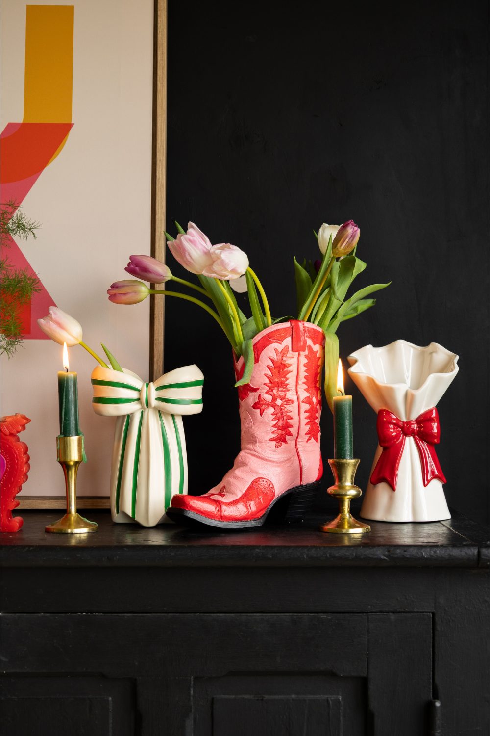 Gold candlesticks and pink and red cowboy boot vase holding tulips on dark wood shelf