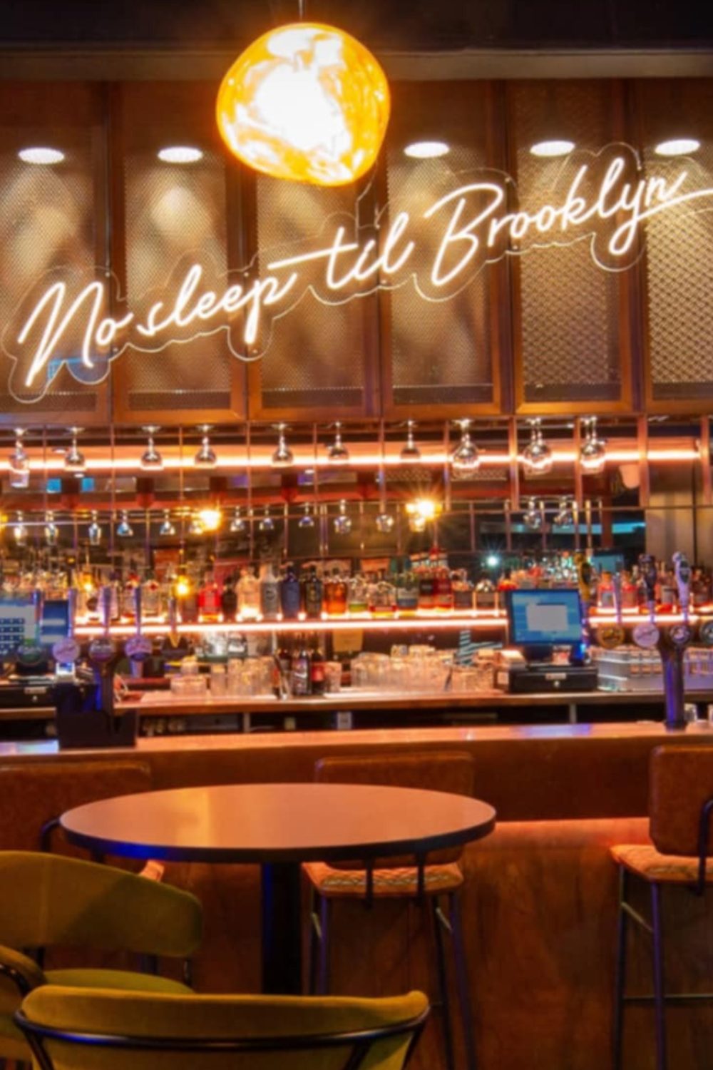 Round table and mustard yellow chairs in front of mirrored bar below white neon sign and round lamp