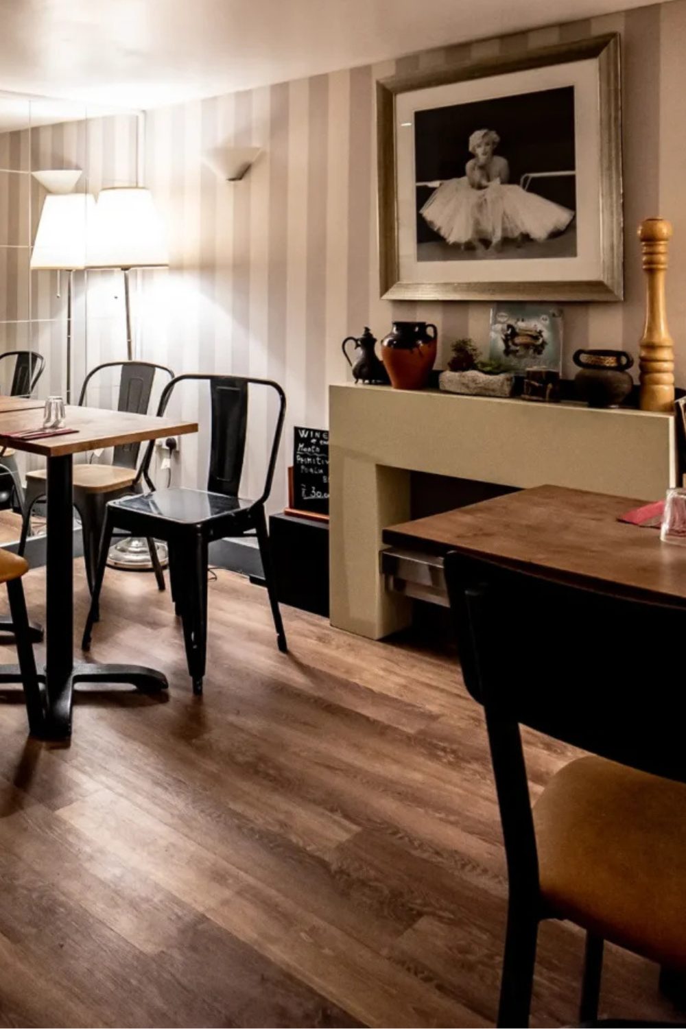 Restaurant interior with white and grey striped wallpaper, wooden floor, white fireplace, metal chairs, and framed portrait of Marilyn Monroe