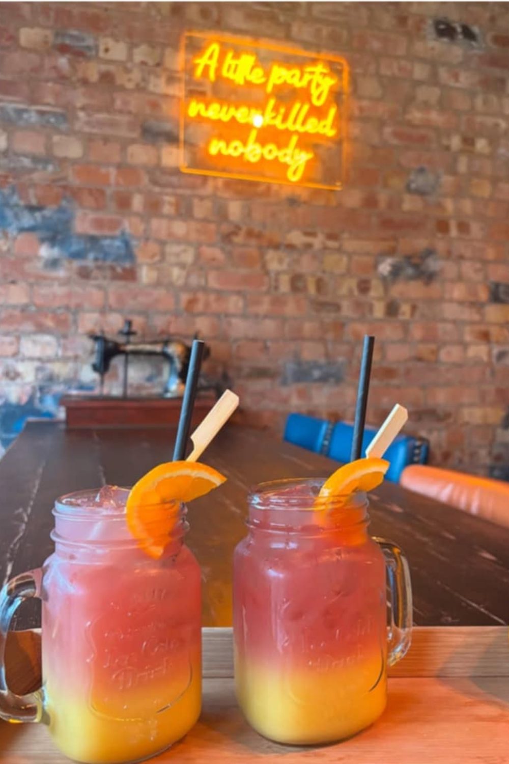 Wooden table with two colourful cocktails in jars in front of brick wall with yellow neon sign