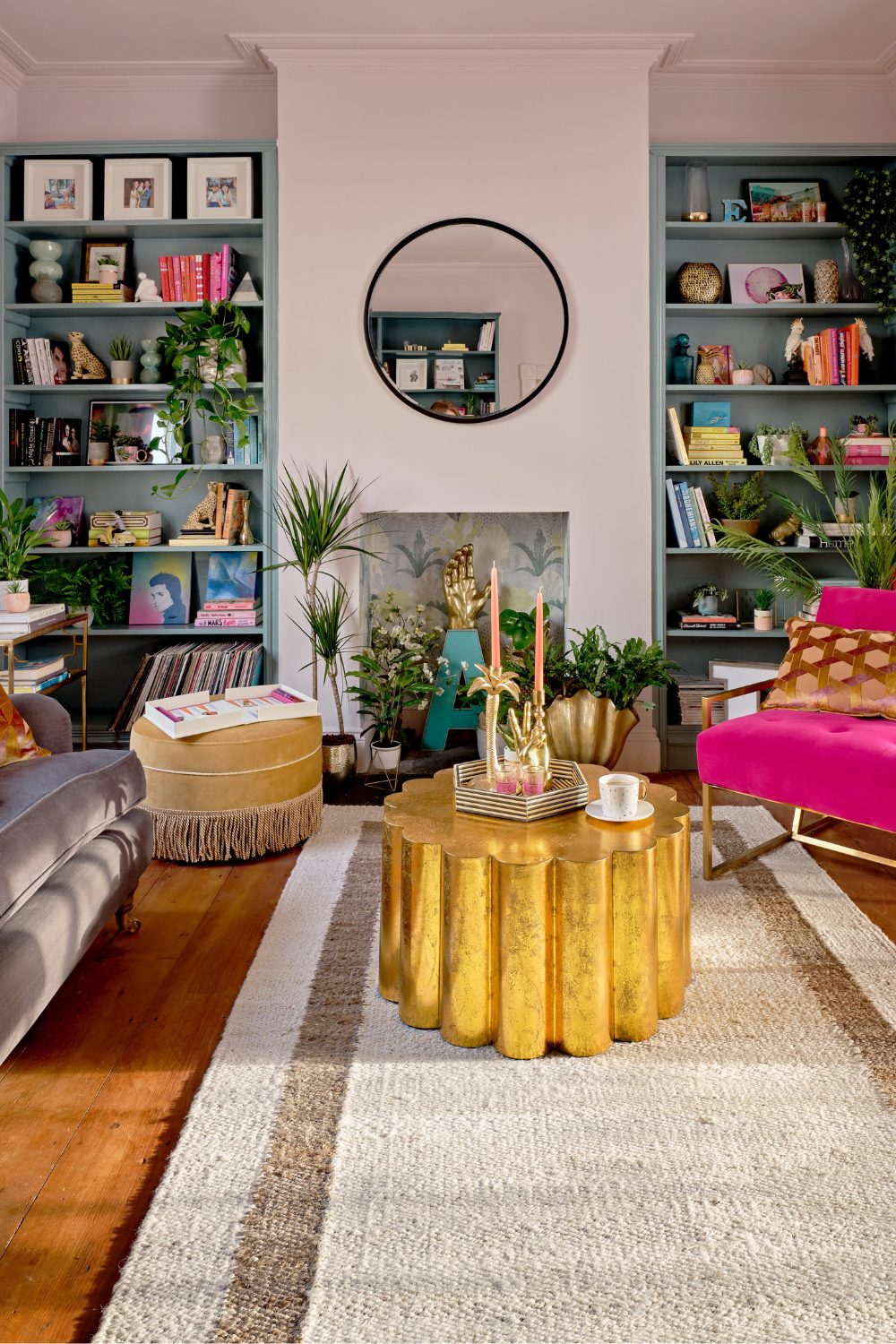 Dracaena plant in gold pot in bright living room with pink armchair, gold scalloped table, blue bookshelves and round mirror