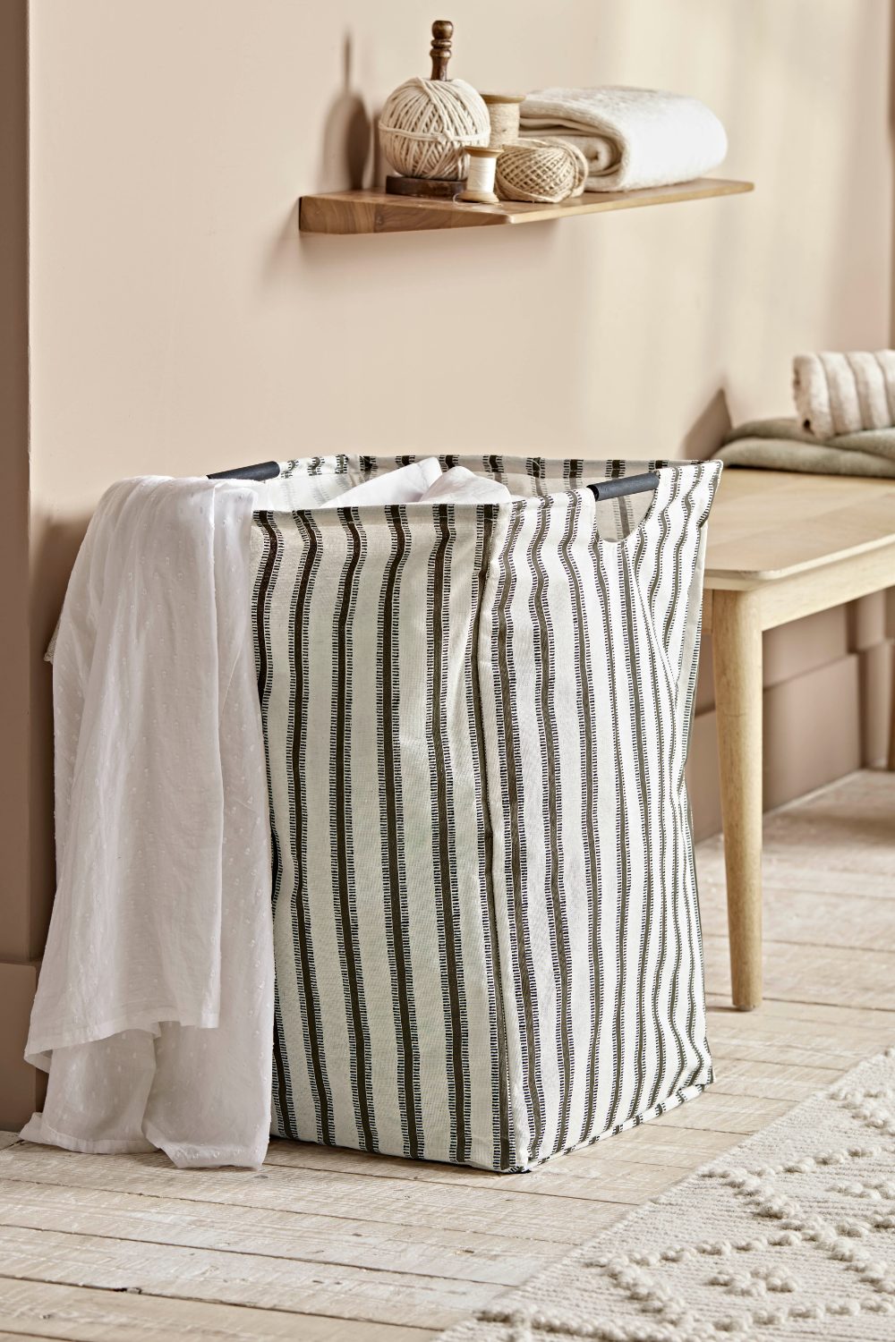 Vertical stripe fabric hamper on wooden floor below wooden shelf holding twine