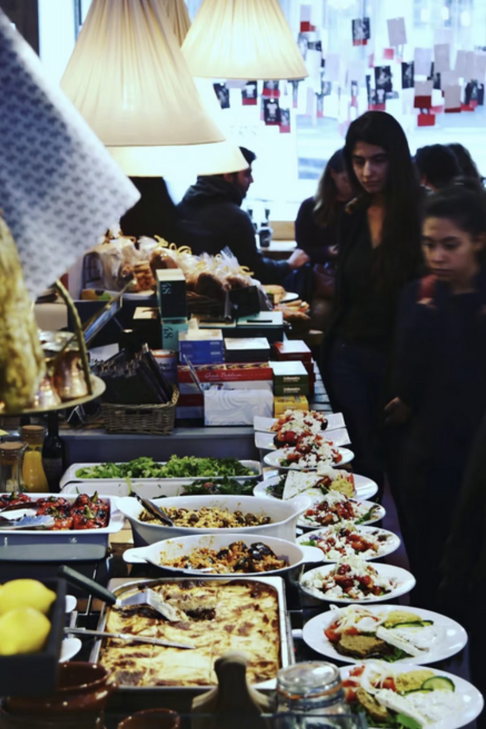 Line of people looking at table filled with plates and dishes of Greek food below collection of white lampshades and hanging cards