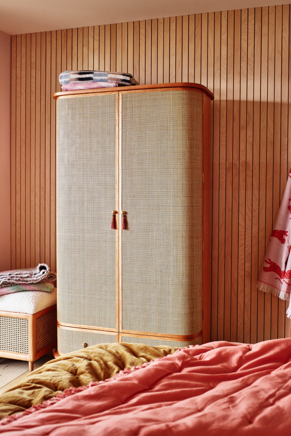 Rattan and wood wardrobe against a slatted wooden wall with bed covered in pink blankets