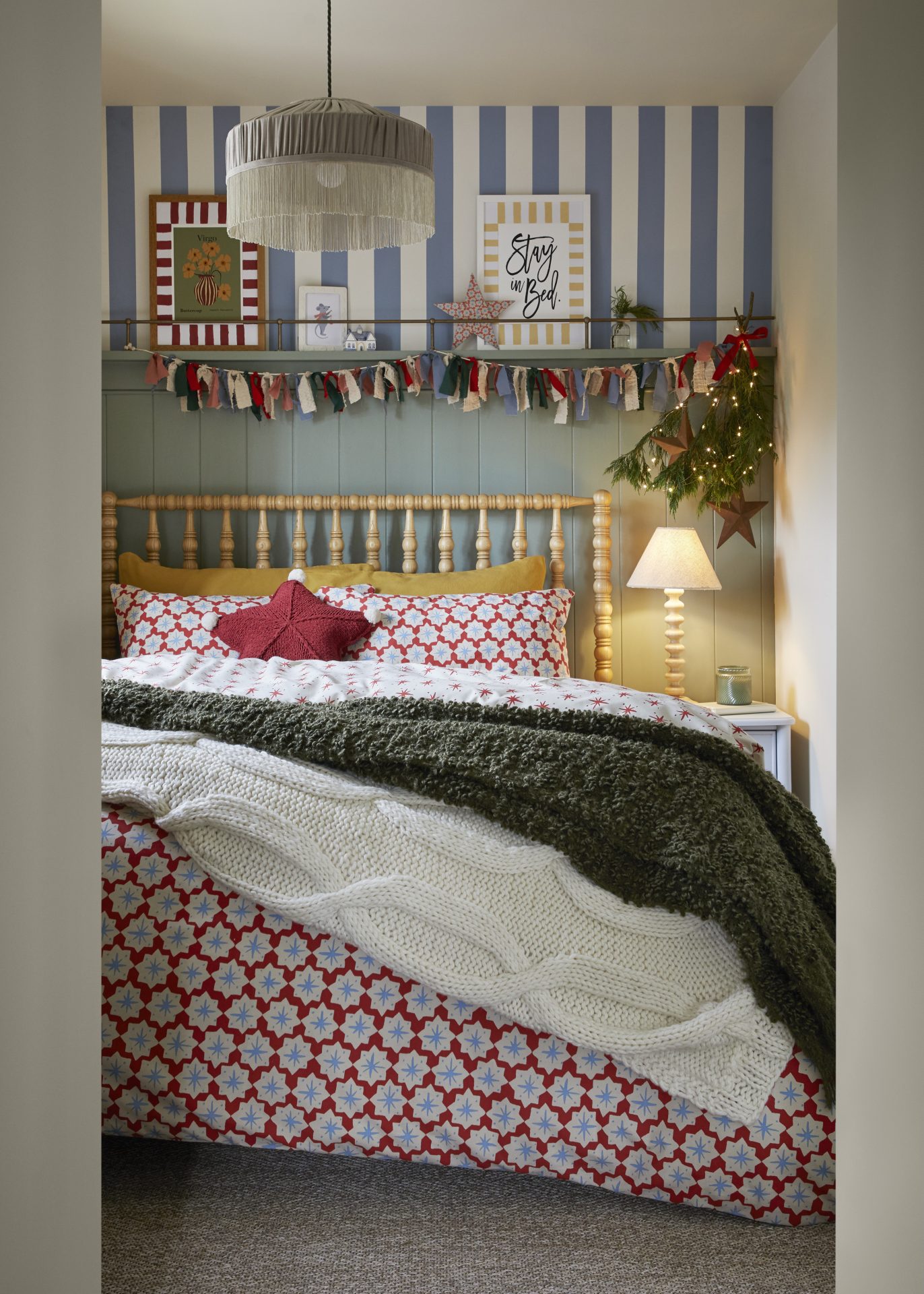 Wooden-framed bed with layers of covers below blue and white striped accent wall and red, white and blue cloth scrap hangings.