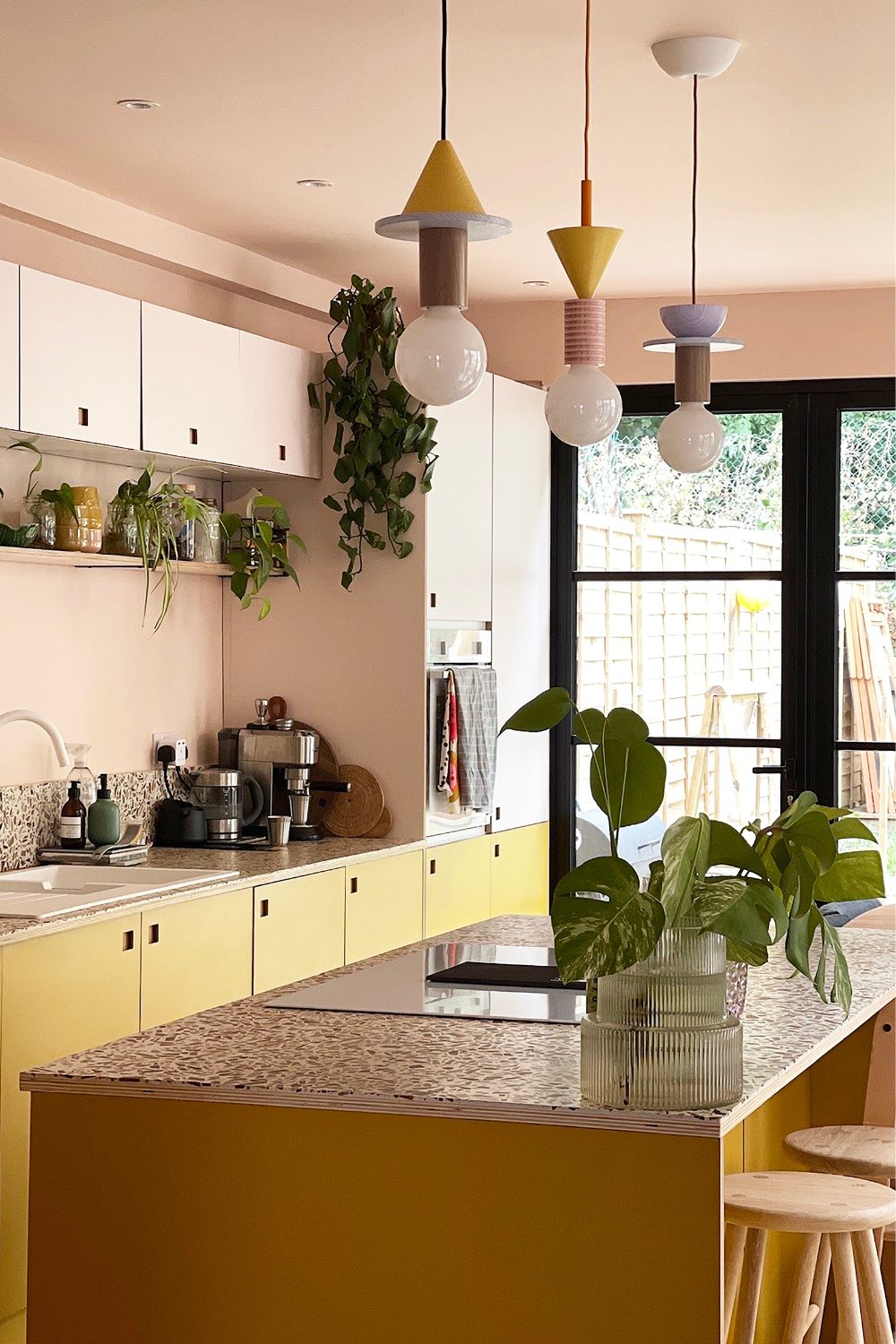 Peach kitchen with yellow cabinets, statement hanging lamps, plants in glass vase, window with garden view and wooden stools
