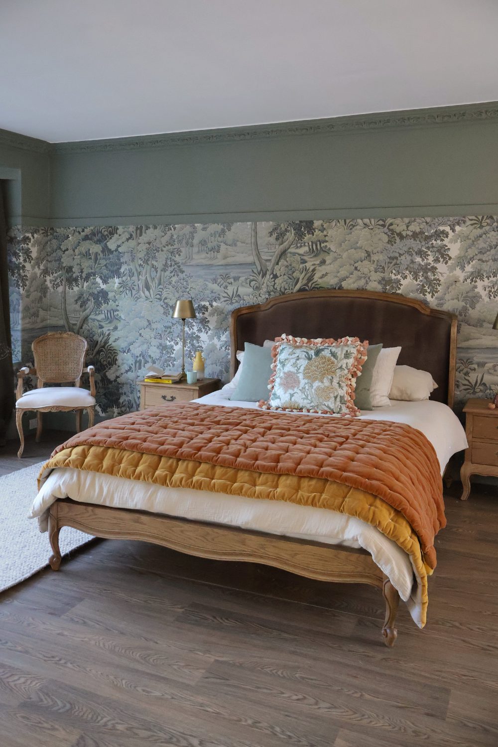 Wooden-framed bed with orange throws on dark wood floor in front of muted green landscape wallpaper