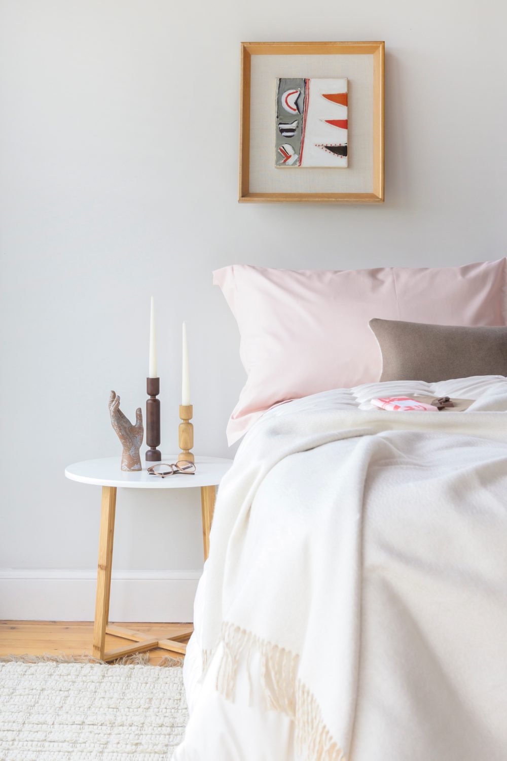 White bedroom with white bedside table holding wooden candlesticks, framed red and white print and bed with pink pillow