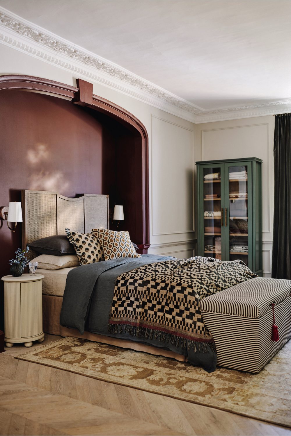 Bed with patterned blanket and striped ottoman in dark red wall recess, next to green glass-fronted cabinet and white bedside table on wooden floor