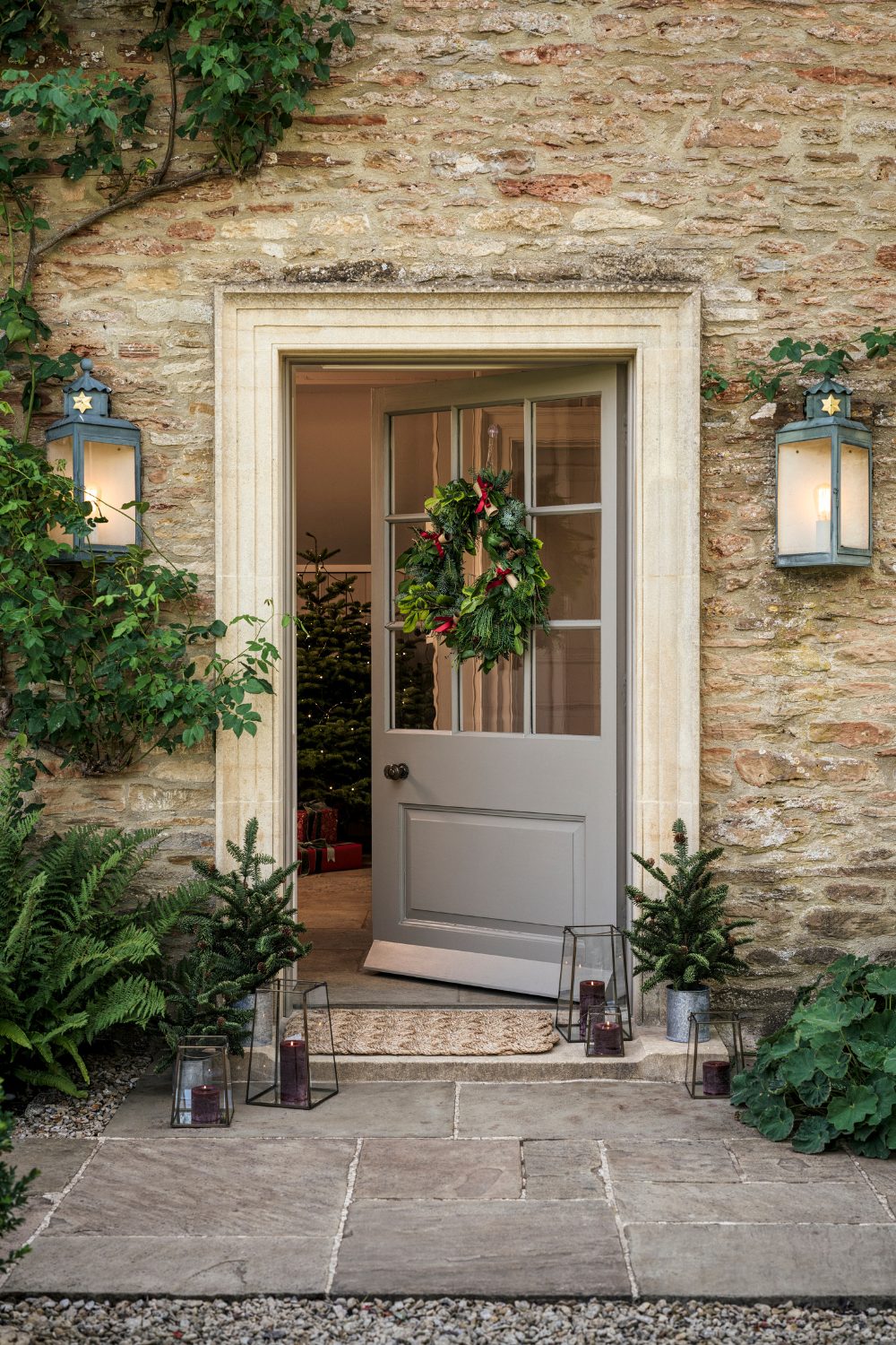 Open grey front door with wreath in stone house flanked by metal lanterns with paving stones in front