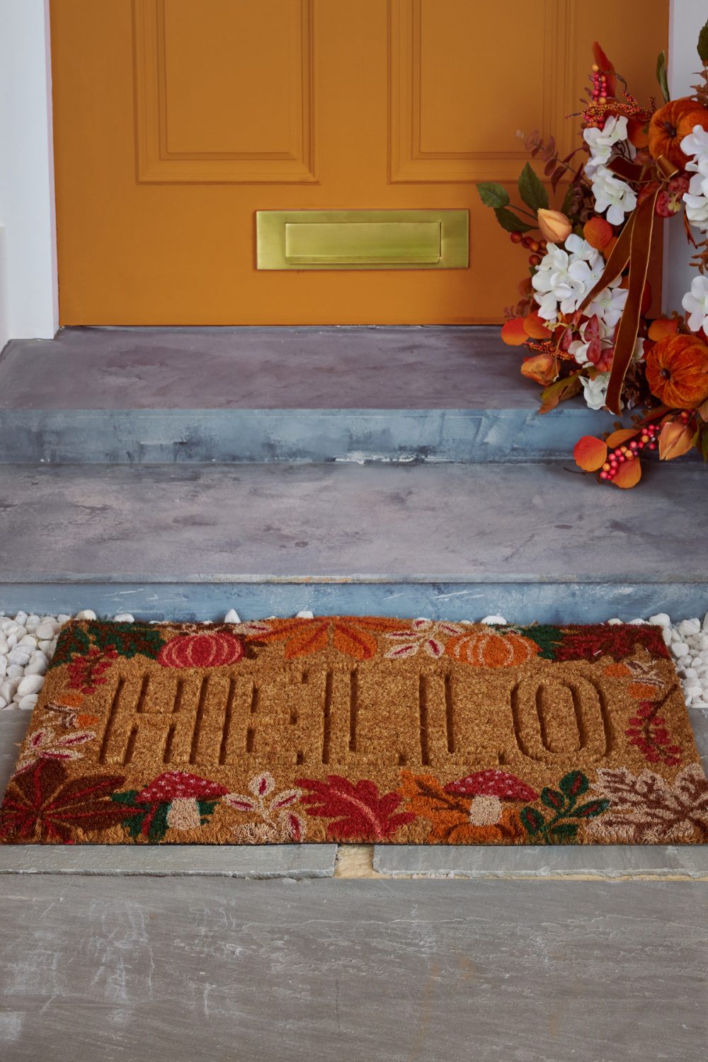 Orange front door with orange wreath and stone steps behind a brown doormat with pumpkin design
