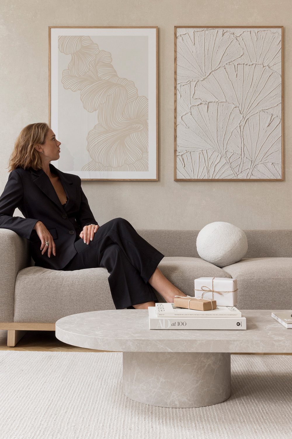 White woman on grey sofa in white living room looking at framed white prints on wall
