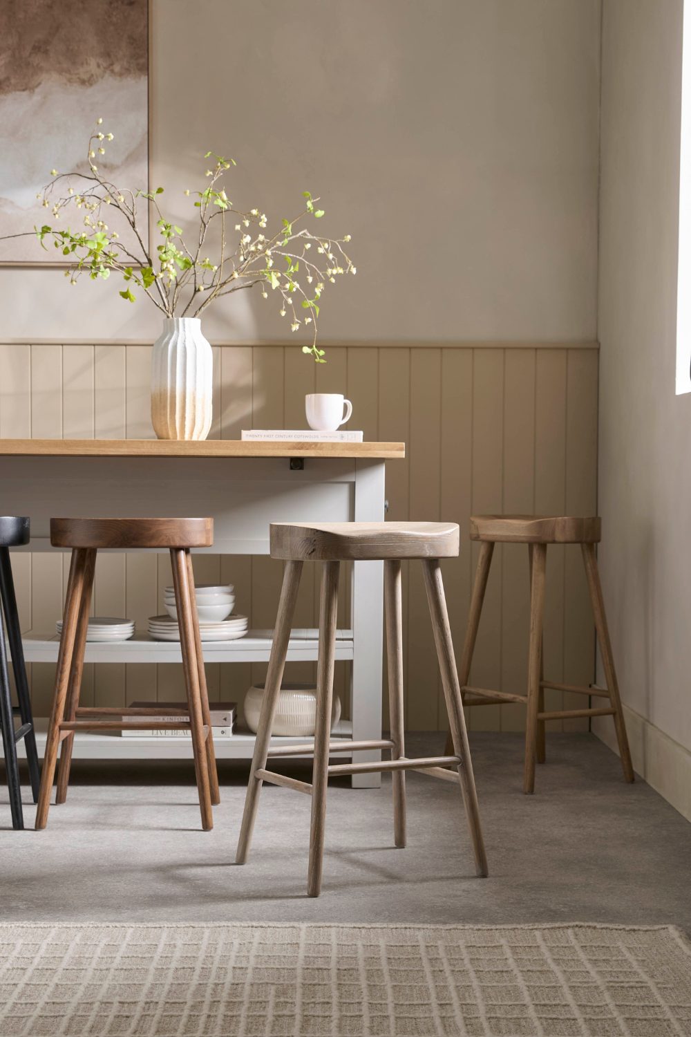 Kitchen in neutral tones with wooden table, flowering branches in white vase, four wooden stools and grey rug
