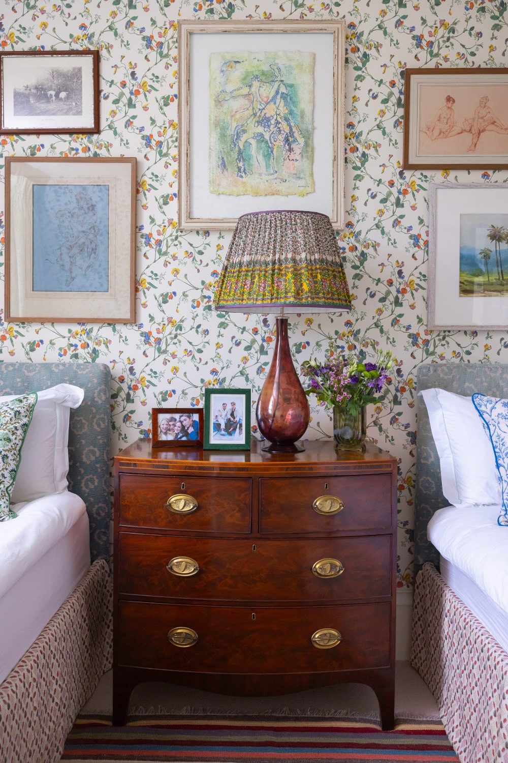 Wooden drawers holding floral lampshade between two beds in front of floral wallpaper with framed drawings
