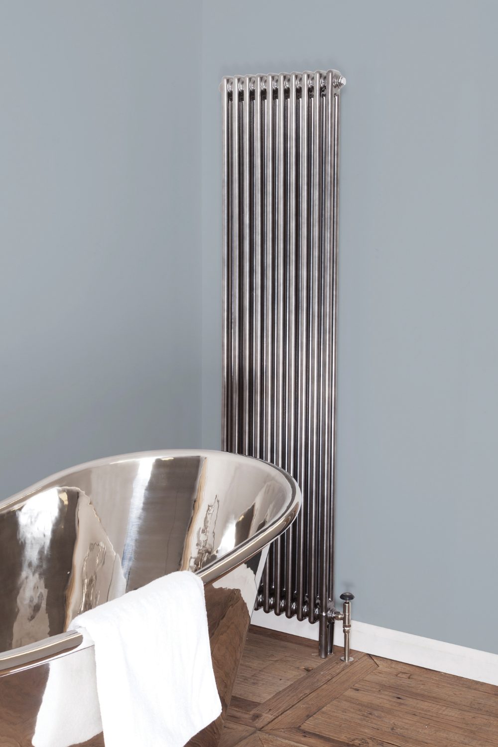Modern vertical silver radiator in grey-blue bathroom behind silver bath