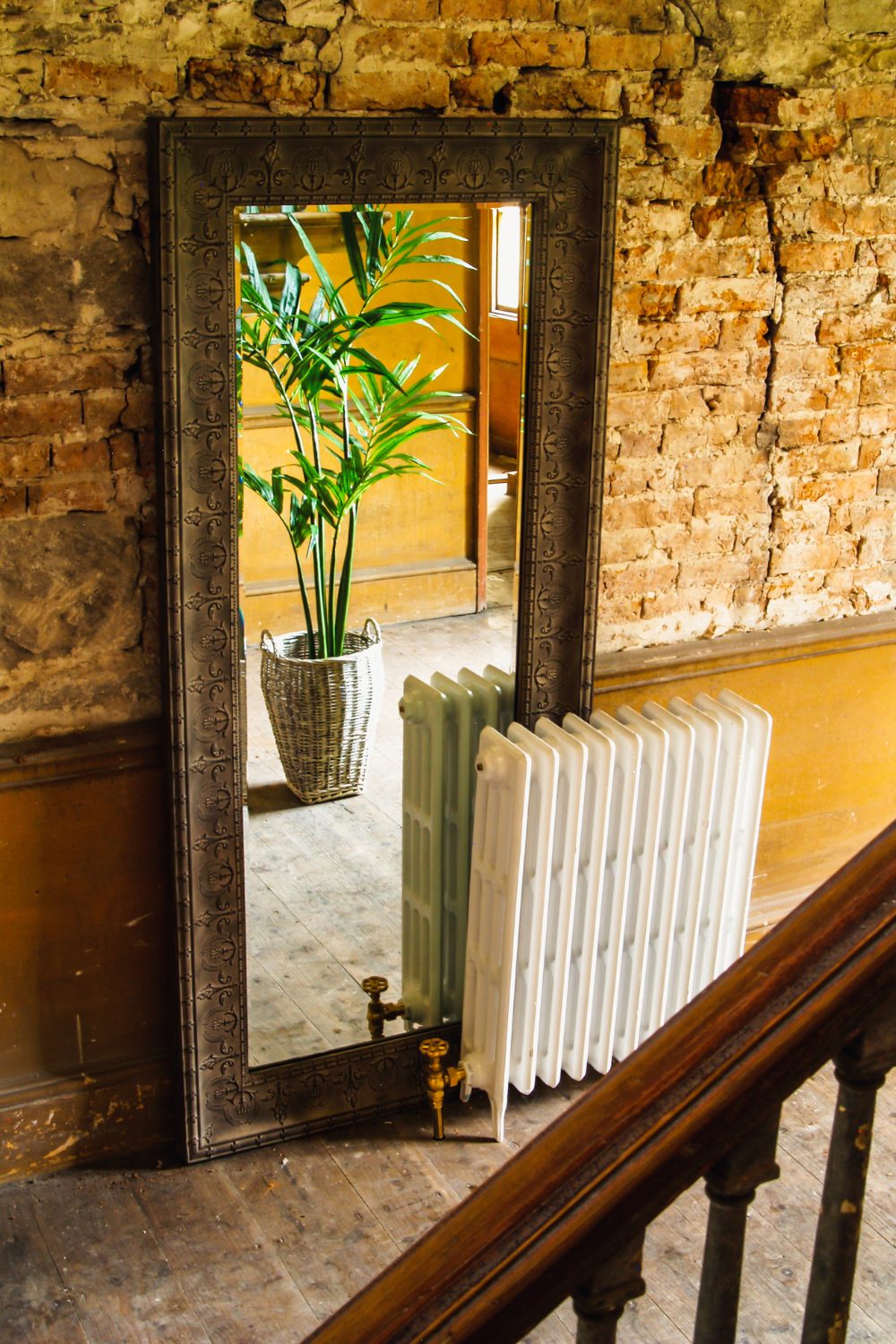 White radiator against a metal-framed mirror reflecting a plant in wicker basket against a distressed brick wall