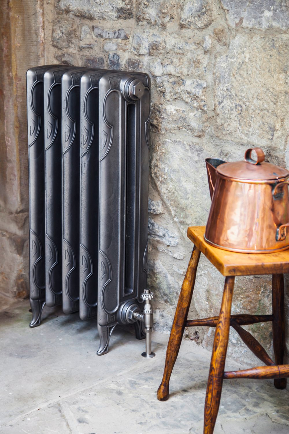 Black metal radiator on stone wall next to copper kettle on wooden stool