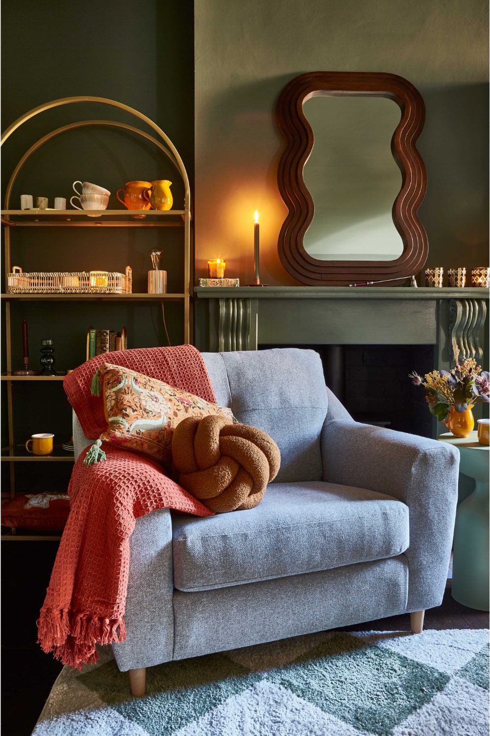 Blue armchair with red throw, orange cushion and brown cushion in front of dark green fireplace, gold metal bookshelf and curved wooden mirror